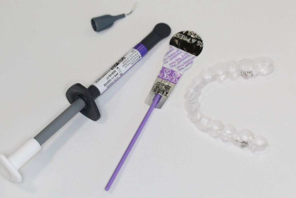 Dental materials including a syringe applicator, a dental brush with a purple handle, a sealed dental capsule, and a clear aligner tray on a white surface.